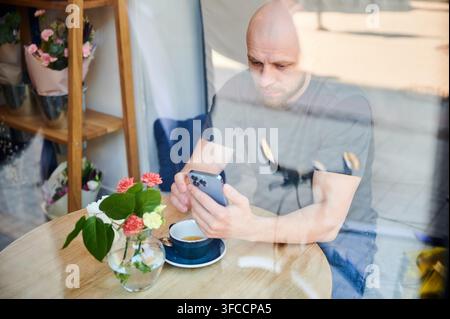 Man using smartphone in cafe seen through window, browsing or messaging at table with coffee and flowers, reflections on glass, modern lifestyle and digital communication concept Stock Photo