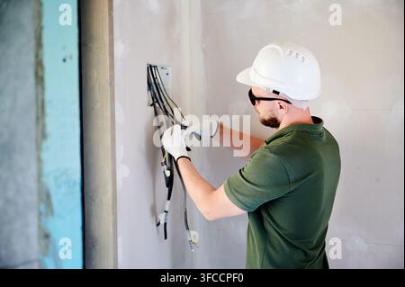 Electrician connecting wires inside wall junction box and organizing cables. Worker in helmet and gloves preparing electrical system for installation, interior construction and wiring setup concept Stock Photo