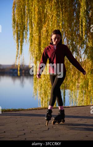 Female skater riding on lakeside trail, relaxed full body view framed by willow trees and fallen leaves. Suitable for leisure, healthy habits, mobility or outdoor editorial use Stock Photo