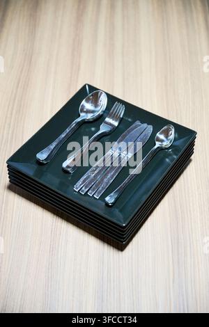 Cutlery set placed on stacked black plates on wooden table. Top view of spoons, forks and knives prepared for dining, modern hospitality, kitchen or rental apartment concept Stock Photo