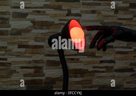 Black robotic or Prosthesis hand reaches toward a glowing yellow bulb in a modern desk lamp against a stone wall, useful for concepts of AI, innovatio Stock Photo