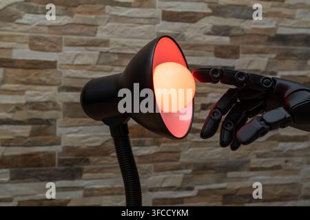Black robotic or Prosthesis hand reaches toward a glowing yellow bulb in a modern desk lamp against a stone wall, useful for concepts of AI, innovatio Stock Photo