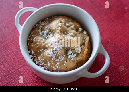 High angle close up of a golden apple cake with caramelized edge and powdered sugar in a white ceramic cocotte on red textured fabric Stock Photo