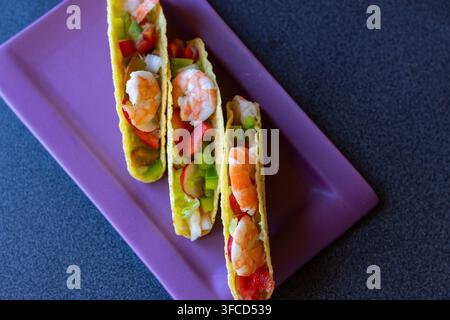 Top view of three crunchy shrimp tacos with radish, lettuce and diced peppers on a purple plate over dark grey with copy space Stock Photo