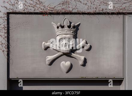 Stone relief sculpture of a crowned skull and crossbones with a small heart mounted on a wall casting a dramatic shadow. Stock Photo