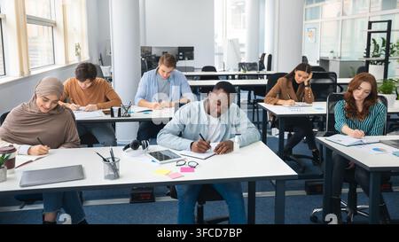Modern Education Concept. Portrait of diverse group of students sitting at desks in classroom at university, taking test, entrance examination or writ Stock Photo