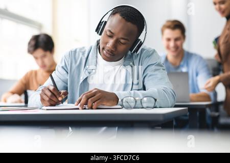 Studying And Learning Concept. Portrait of focused young black man in wireless headset sitting at table in lecture hall in highschool, writing in note Stock Photo