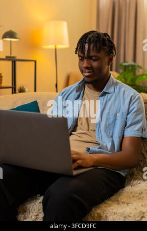 African American young man on home sofa works on laptop checking text and fixing small details with focused attention. Black guy on home sofa edits a document and reviews work with calm concentration. Stock Photo