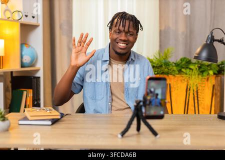 African American young man on home table records short online lesson on smartphone and speaks with confidence at home. Black guy on home table creates useful online lesson and shares advice indoors. Stock Photo
