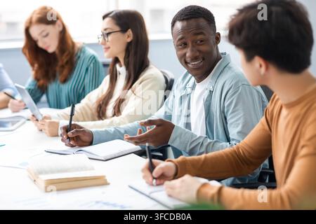 Cheerful male colleagues enjoying working process and cooperation, writing in notebooks and talking, skilled students preparing homework task together Stock Photo