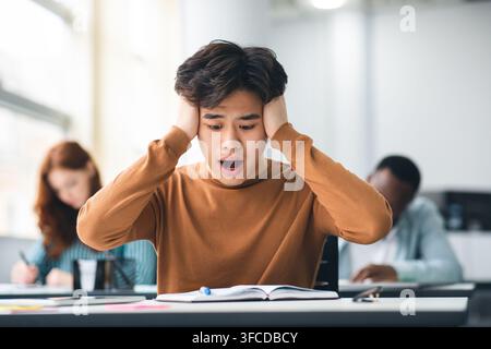 Portrait of shocked asian freshman sitting at table in classroom, grabbing clasping his head in horror and panic, looking at hard exam questions or te Stock Photo