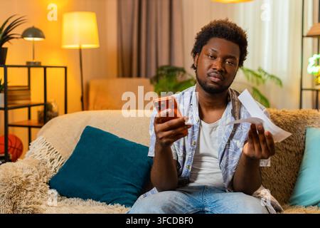 African American young man on home sofa holds bill checks amount and looks concerned with smartphone. Black guy on home sofa feels surprise and starts counting monthly expenses carefully in phone app. Stock Photo
