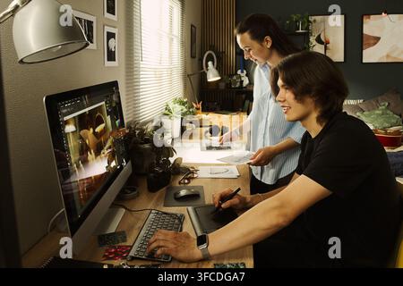 Young multiethnic couple of designers working on computer in home office, editing photos using graphics tablet. Perfect for remote work and creative agency marketing Stock Photo