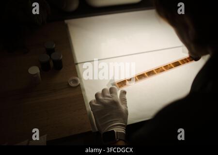 Over shoulder view of photographer wearing protective glove, examining analog film strip on light table. Image represents photography archiving and retro media services Stock Photo