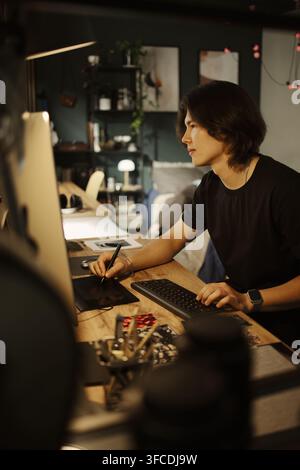 Vertical image of young Asian man working on computer using graphics tablet at home office, illustrating remote work and digital design career concepts for creative agency marketing Stock Photo