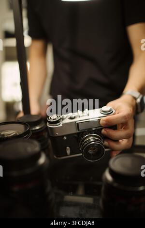 Young man holding vintage film camera near modern lenses, preparing equipment for photoshoot. Image suitable for photography school promotion or camera store advertising, vertical Stock Photo