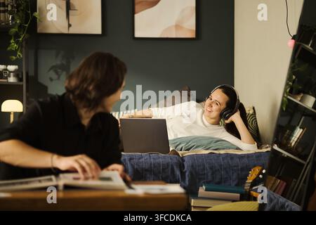 Young couple studying together in bedroom, one lying on bed with laptop and headphones while other reads book, representing remote learning and student lifestyle Stock Photo