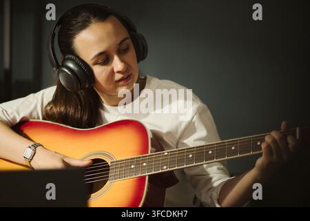 Young Caucasian woman wearing headphones playing acoustic guitar during online music lesson. Image represents digital learning and is suitable for e-learning platform marketing Stock Photo