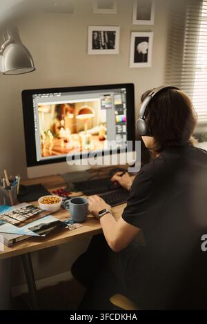 Rear view of young Asian man wearing headphones, editing photo on computer using graphics tablet at home office. Image suitable for remote work and digital creator campaigns, vertical Stock Photo