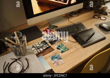 Graphic designer workspace featuring computer monitor, keyboard, graphics tablet, and printed reference photos on wooden desk, representing creative workflow, high angle shot Stock Photo