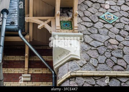 A carved stone bracket bearing the year 1904 and the signature "C. Pain" supports a wooden balcony overhang on a façade on Rue Félix Faure in Nancy, Grand Est, France, with ceramic tile medallions decorating the rubble-stone wall beside it. Stock Photo