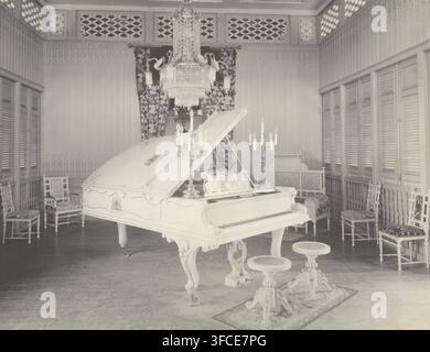 White wing for the Sultan of Langkat, photograph, inscription: 'gelieft von Schiedmayer & Söhne Stuttgart ein Auftrag von Sultan von Langkat, durch die Firma P. Sandel Tandjong Poera', check stamp: 'CJ Kleingrothe, Medan - Sumatra', photographer: Carl J. Kleingrothe, Tandjong Poera, 1885 - 1914, Sumatra, North Sumatra, paper, platinum process, height 284 mm x width 364 mm, grand piano, Medan., A pale grand piano with an ornate carved body and open lid sits centrally in a wood-paneled room, flanked by decorative candelabras on its surface and a pair of carved round stools with detailed pedestal Stock Photo