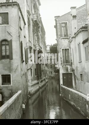 Film negative with shot of Venice, negative, photograph, Venice, c. 1907 - c. 1935, photographer, plastic, organic material, cellulose nitrate film, gelatin silver negative, gelatin silver negative on cellulose nitrate film, height 90 mm x width 120 mm., A narrow canal runs between tall, weathered buildings with worn facades, shuttered and arched windows, and small balconies; the canal water reflects light and appears calm, bordered by brick and stone walls and a low bridge or railing in the foreground, with a few doorways opening onto the water, visible pipes and wires strung along the Stock Photo