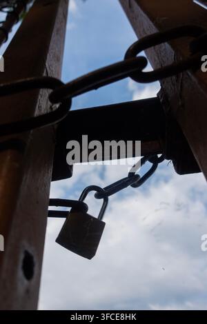 Close-up of a padlock and heavy chain securing weathered wooden boards, viewed from below against a blue cloudy sky Stock Photo