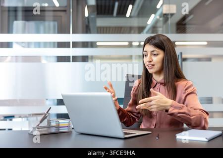 Young businesswoman actively participating in a video conference from her modern office, communicating and explaining concepts while using a laptop for remote work Stock Photo