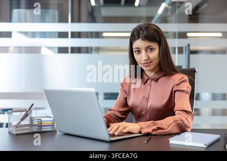 Young professional woman in a modern office sits at a desk typing on a laptop, looking confidently at the camera with a focused, determined expression during daytime work Stock Photo