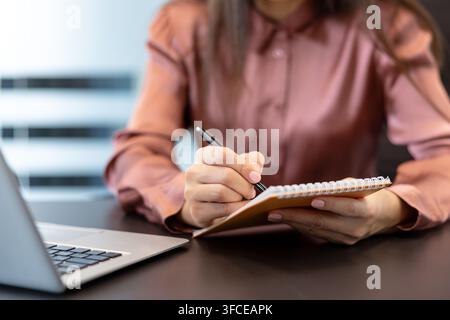 Woman's hands holding a spiral notepad and writing with a pen, capturing ideas, taking notes, or making a plan while working at a desk with a laptop in a modern office environment Stock Photo