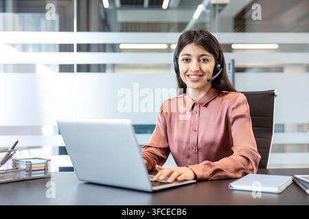 Woman in pink blouse wearing a headset, smiling at camera while typing on laptop at office desk, providing friendly customer support and virtual assistance during workday Stock Photo