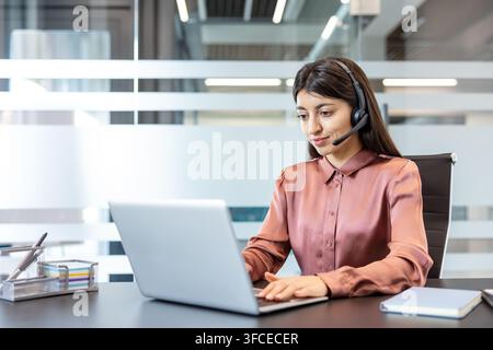 Young female customer service representative with headset typing on a laptop in a modern office, providing online technical support and virtual assistance with a confident smile Stock Photo