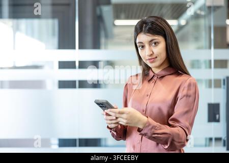 Young businesswoman with long dark hair stands in a modern office holding a smartphone, looking at camera with confident, professional expression conveying tech-savvy connection and success Stock Photo