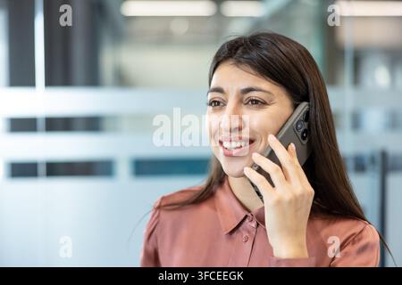 Businesswoman smiling and talking on a smartphone during a professional call in a modern office, conveying confident, friendly communication and successful business interaction Stock Photo
