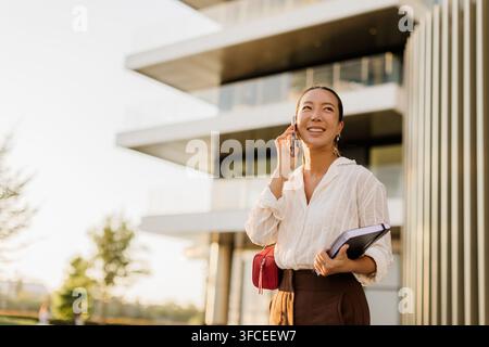 A joyful Korean woman talks on her phone while standing outside a modern building in a sunny park setting. Her cheerful smile brightens the scene. Stock Photo
