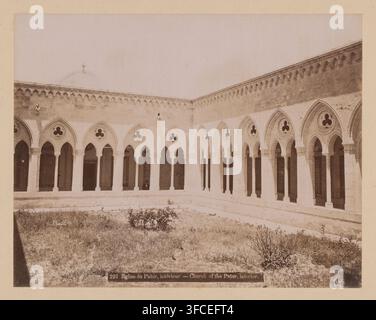 A sepia-toned photograph shows an inner courtyard with a covered arcade of pointed arches resting on columns, each arch containing a circular quatrefoil opening above the archway; the arcade forms two sides of a rectangular cloister with a low stone wall and a grassy courtyard in front, scattered with small plants and a few bushes; above the arches a stone frieze with repeated decorative motifs runs along the top of the walls, and a domed roof with a small finial is faintly visible behind the wall on the left; the scene is empty of people and has an overall aged, weathered appearance. Stock Photo