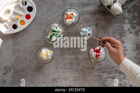 Overhead view of a hand painting decorative Easter eggs surrounded by colorful eggs and a paint palette on a gray textured background Stock Photo