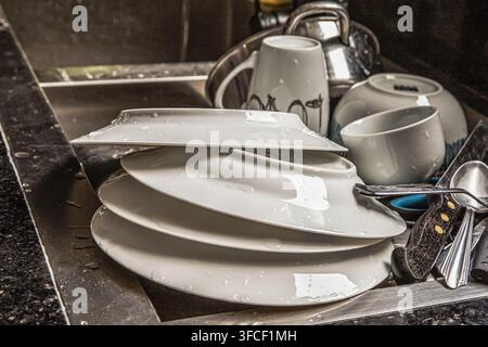 Wet plates and cups stacked in a stainless steel kitchen sink Stock Photo
