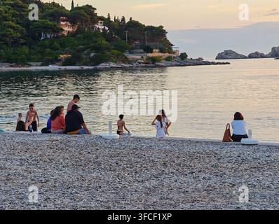 People sitting on the pebbly shore of Lapad Beach, enjoying a serene sunset view over the tranquil Adriatic Sea. Stock Photo