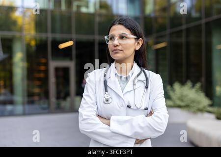 Young indian woman doctor in lab coat with stethoscope and glasses, arms crossed and thoughtful, standing outside a modern clinic building, confident and professional portrait Stock Photo