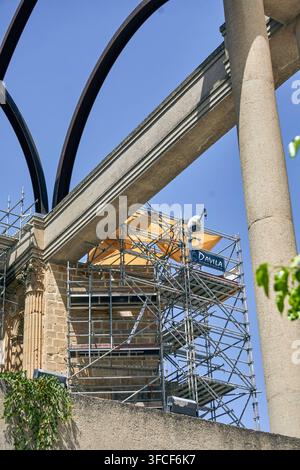 Construction worker on scaffolding beside a historic stone facade, with large columns and curved metal structures under blue sky in Baeza Stock Photo