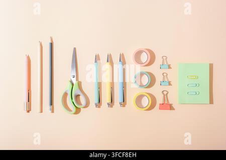 Pastel stationery supplies arranged in a neat flat lay on a peach background, featuring pencils, scissors, pens, tape and clips casting strong shadows Stock Photo