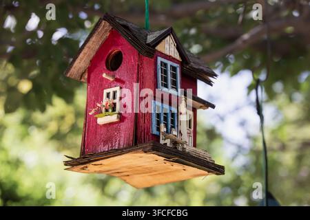 A vibrant, handcrafted birdhouse with red and blue accents hangs from a tree branch, set against a lush garden backdrop in Cramahe. Ideal for themes o Stock Photo