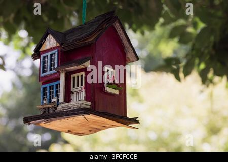 A vibrant, handcrafted birdhouse with red and blue accents hangs from a tree branch, set against a lush garden backdrop in Cramahe. Ideal for themes o Stock Photo