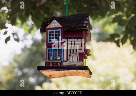 A vibrant, handcrafted birdhouse with red and blue accents hangs from a tree branch, set against a lush garden backdrop in Cramahe. Ideal for themes o Stock Photo