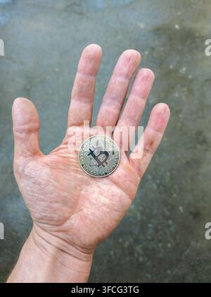 Close-up shot of a silver Bitcoin coin resting in an open palm. The hand is visible from wrist to fingertips, with a blurred textured background. Stock Photo