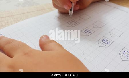 Close up top view of child hand holding pen drawing geometric shapes and houses in grid notebook paper during preschool activity horizontal stock phot Stock Photo