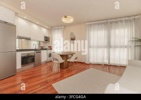 Open kitchen with white cabinets, stainless appliances, round wooden dining table, gray rug, and sheer curtains Stock Photo