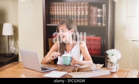 Cheerful young woman with glasses smiles at her laptop while holding a coffee mug during a relaxed break at a wooden home office desk. Stock Photo
